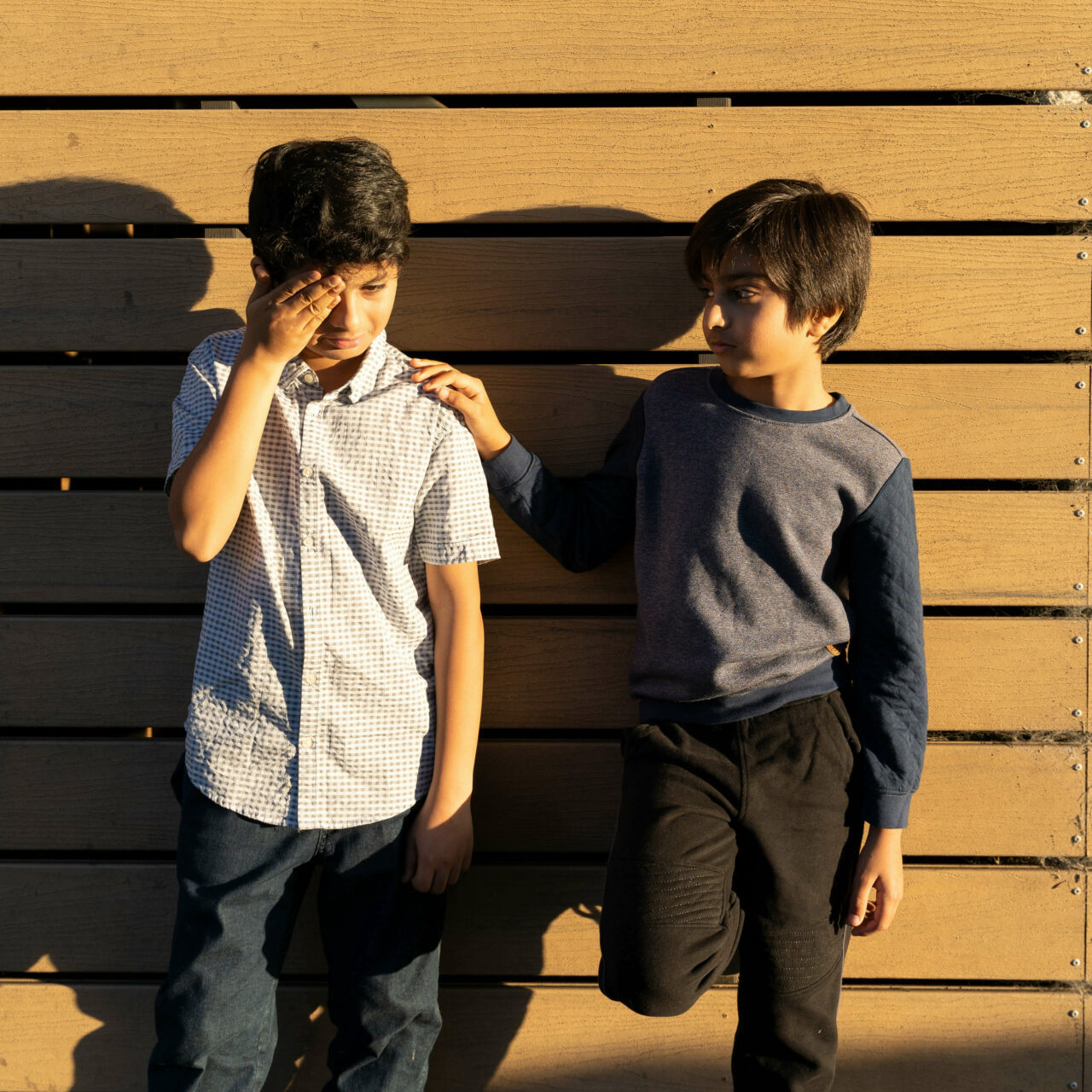 two boys standing against wall, one cries, the other puts his hand on the crying boy's shoulder