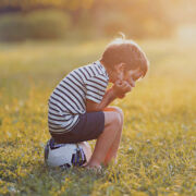 Boy sitting on soccer ball with his head in his hands with sunset in the distance