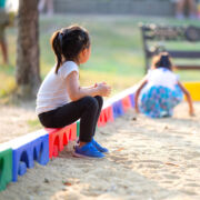 Niña sentada sola en el borde de la caja de arena viendo jugar a otros niños