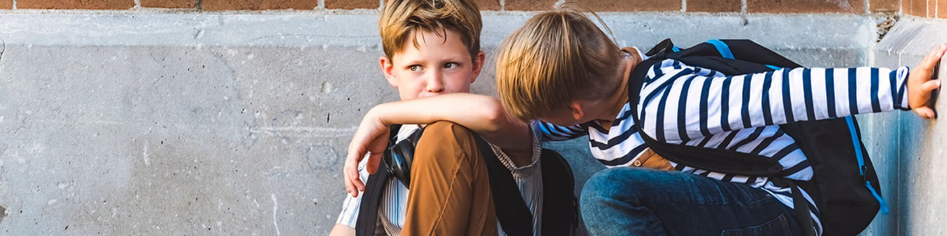 two boys sit on ground, one has his arm around the other who is upset