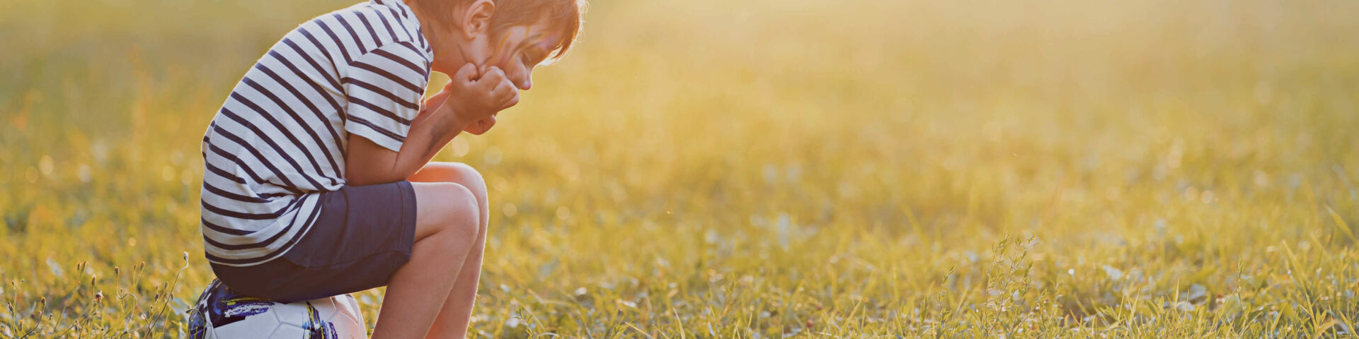 Boy sitting on soccer ball with his head in his hands with sunset in the distance