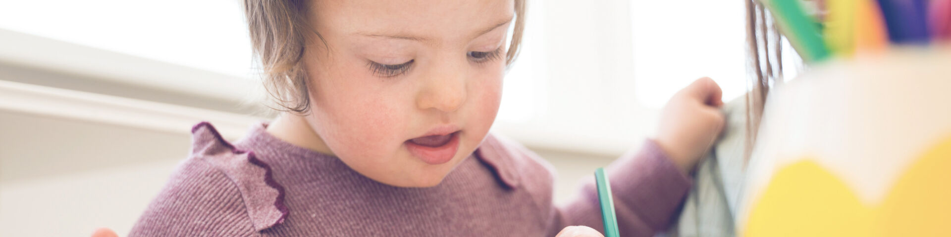 Niño en edad preescolar con síndrome de Down mira el papel mientras sostiene lápices de colores y la maestra dibuja en el papel