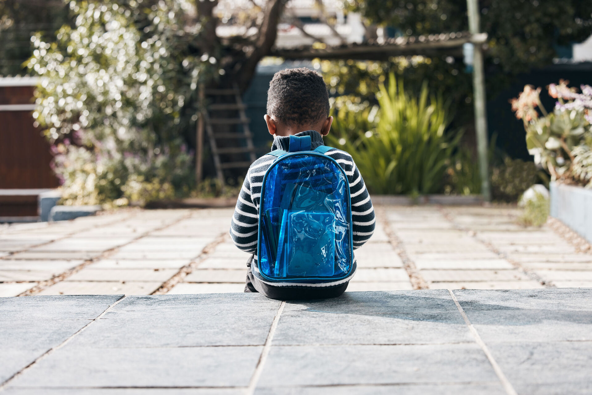 Little boy seated on exterior stone ground, wearing a blue backpack and facing away from the camera.