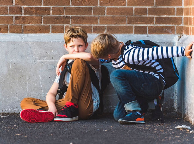 two boys sit on ground, one has his arm around the other who is upset