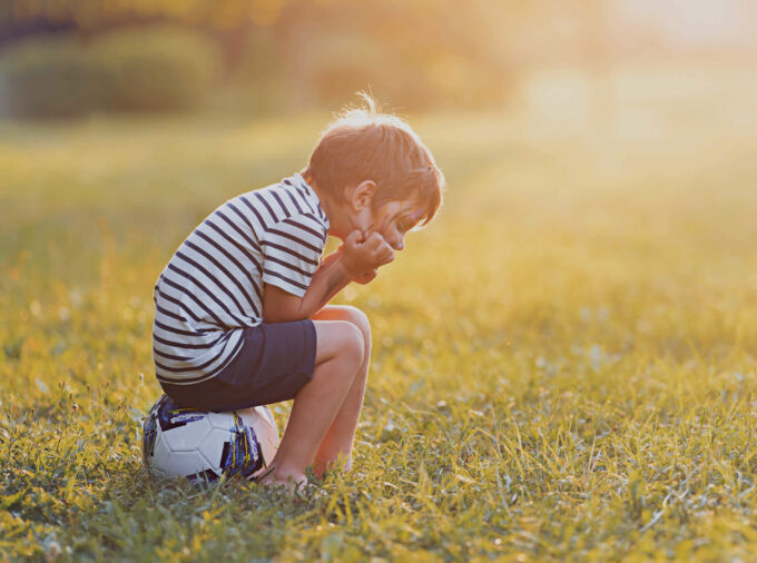 Niño sentado en la pelota de fútbol con la cabeza en las manos con la puesta del sol en la distancia