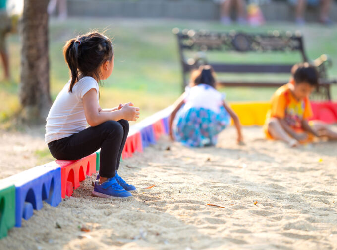 Little girl sitting alone on edge of sandbox watching other kids play