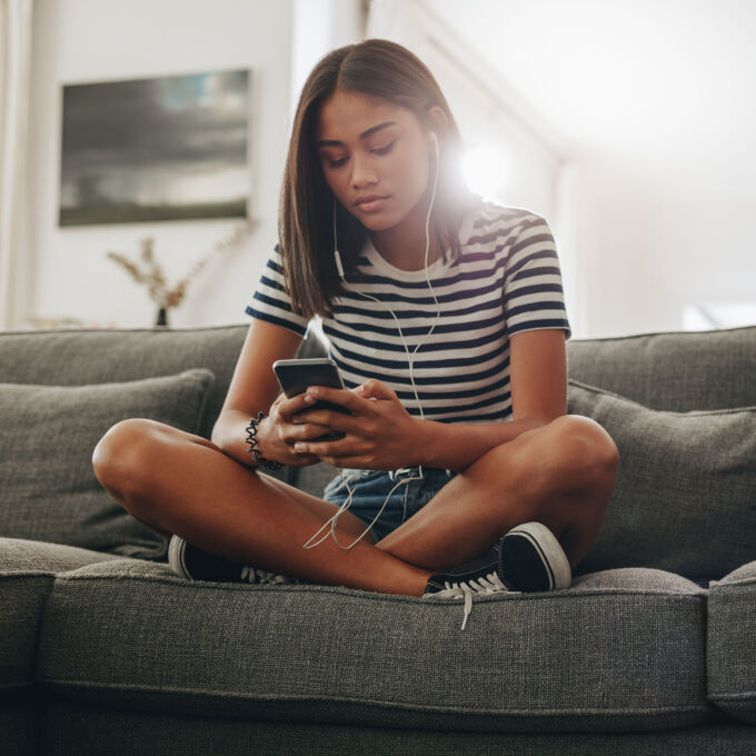 Girl sitting on couch holding phone with headpones on.