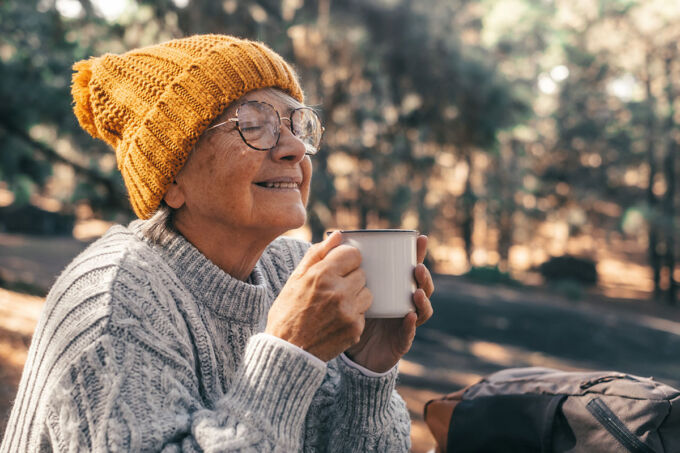 Mujer mayor con gorro de punto y suéter, que se ve tranquila y sentada en un bosque con una taza de café.